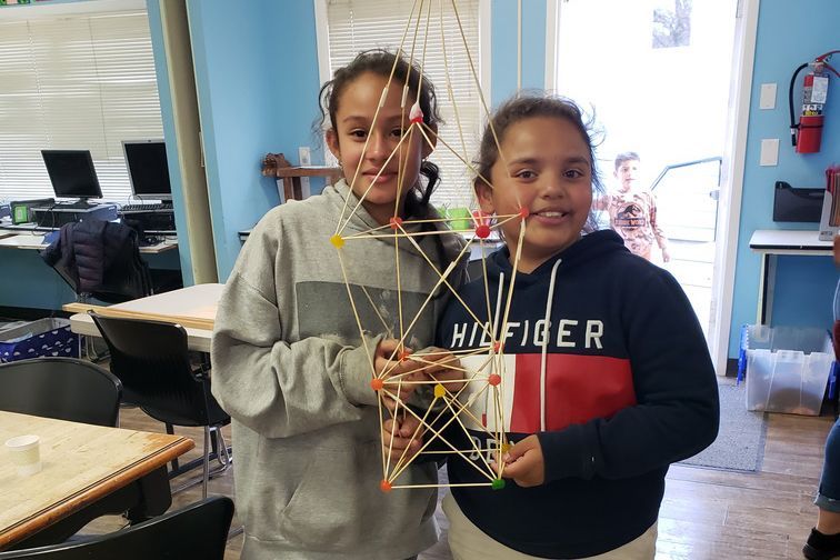 Two girls smile, holding a toothpick and marshmallow tower they built in a classroom.