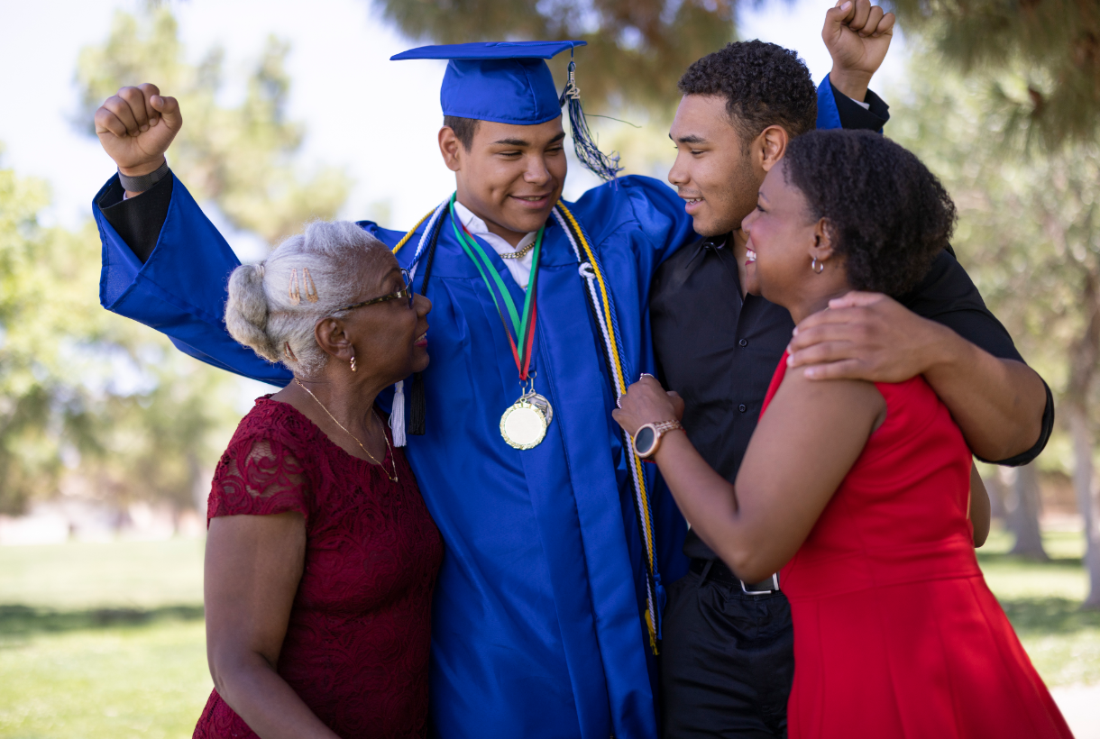 Graduating young man in blue gown celebrates with family outdoors; hugs, joy.