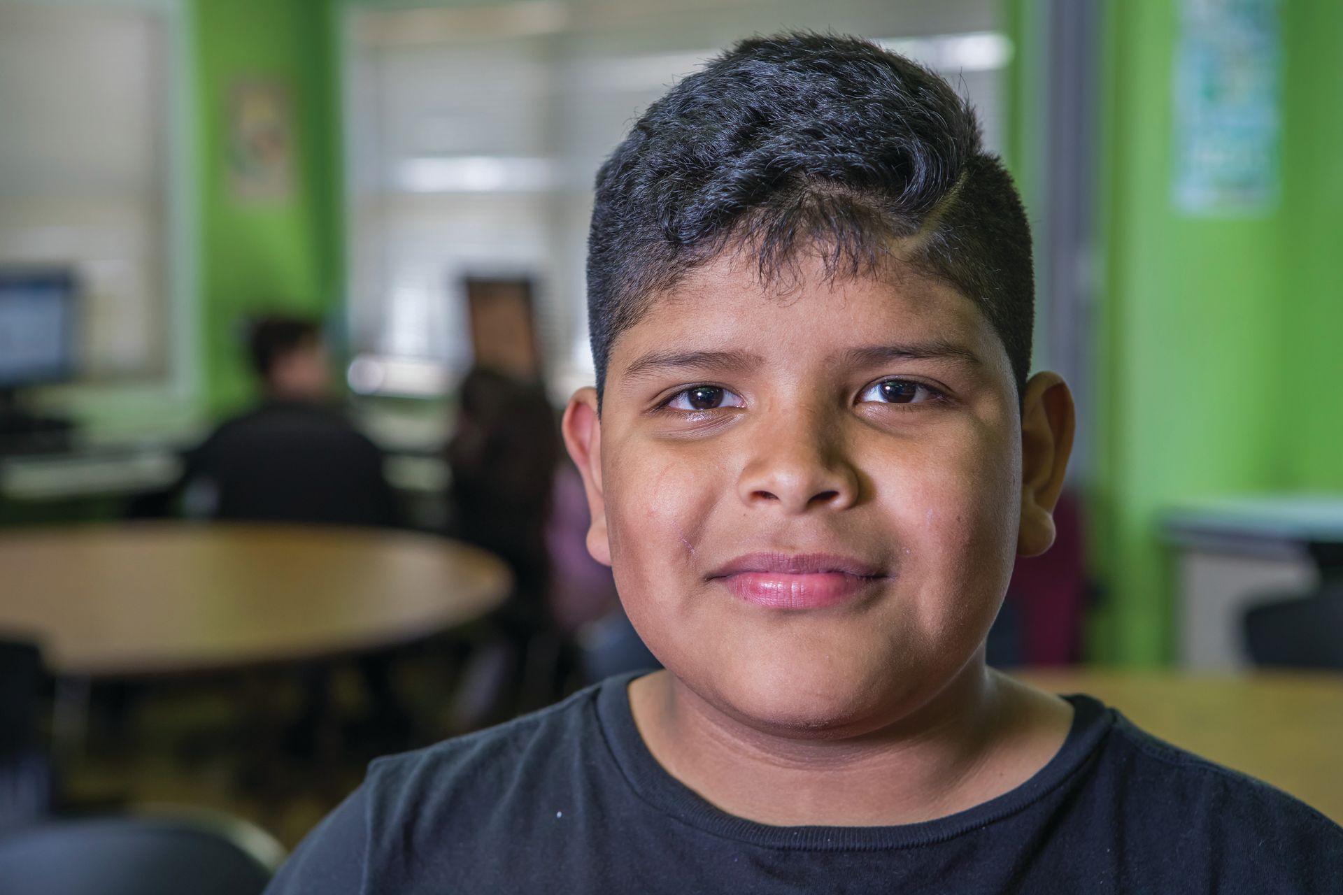 Boy with dark hair and a slight smile in a classroom with green walls.
