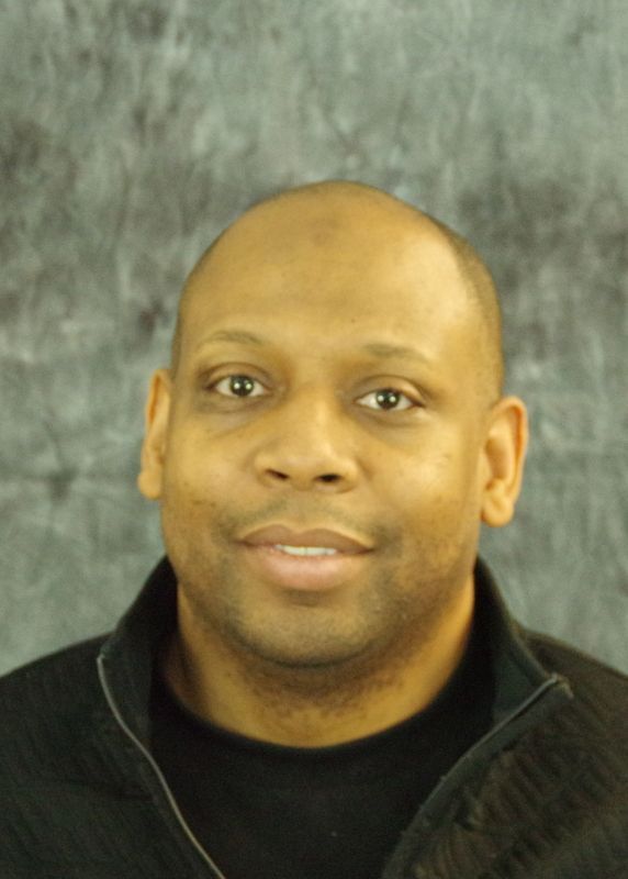 A Black man with a shaved head, smiling, wearing a black sweater. Against a gray background.