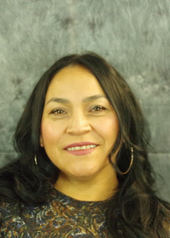 Woman with long dark hair, smiling, wearing hoop earrings, against a mottled gray background.