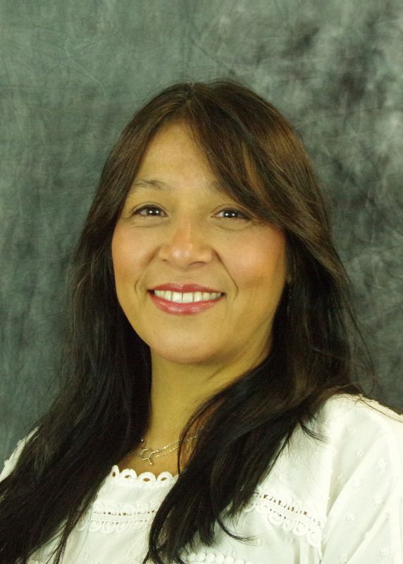 Woman with long dark hair smiles at the camera, wearing a white top, in front of a mottled gray backdrop.
