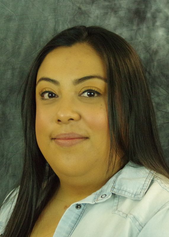 Woman with long dark hair and light blue shirt smiles at the camera. Against a mottled gray background.