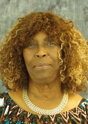 Woman with curly brown hair, wearing a patterned top and a beaded necklace, smiles at the camera.