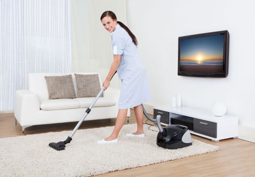 Woman vacuuming a white rug in a bright living room with a sofa and wall-mounted TV