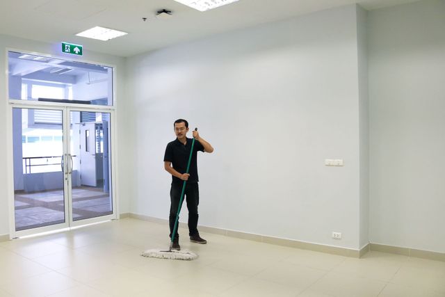 Man holding a mop in a bright empty room with glass doors and white walls.