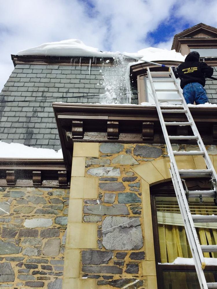 Worker on ladder clearing snow from a slate roof above a stone building.