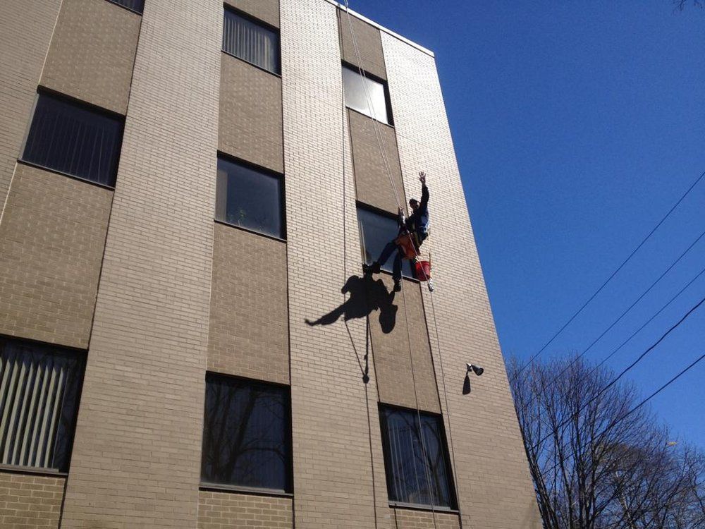 Two window washers suspended by ropes on a tan apartment building against a blue sky