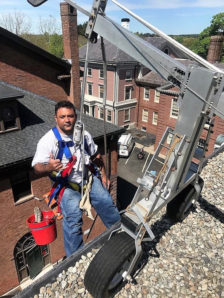 Worker seated in a rooftop harness beside a crane arm, with brick buildings and street below