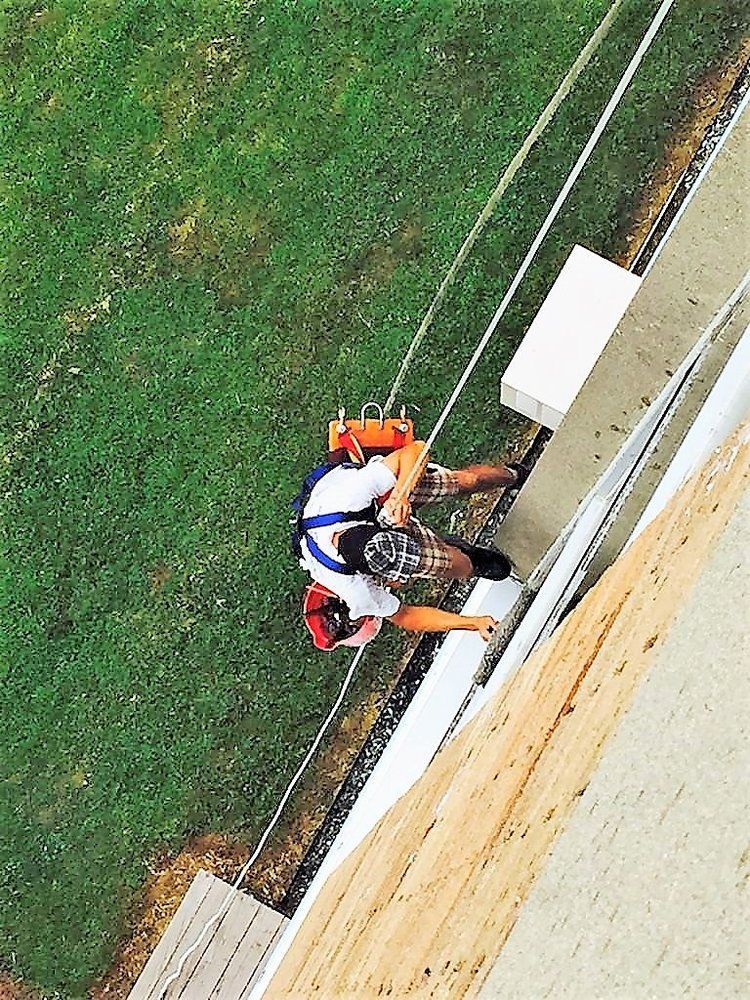 Worker in a safety harness rappelling down a building wall beside a grassy area.