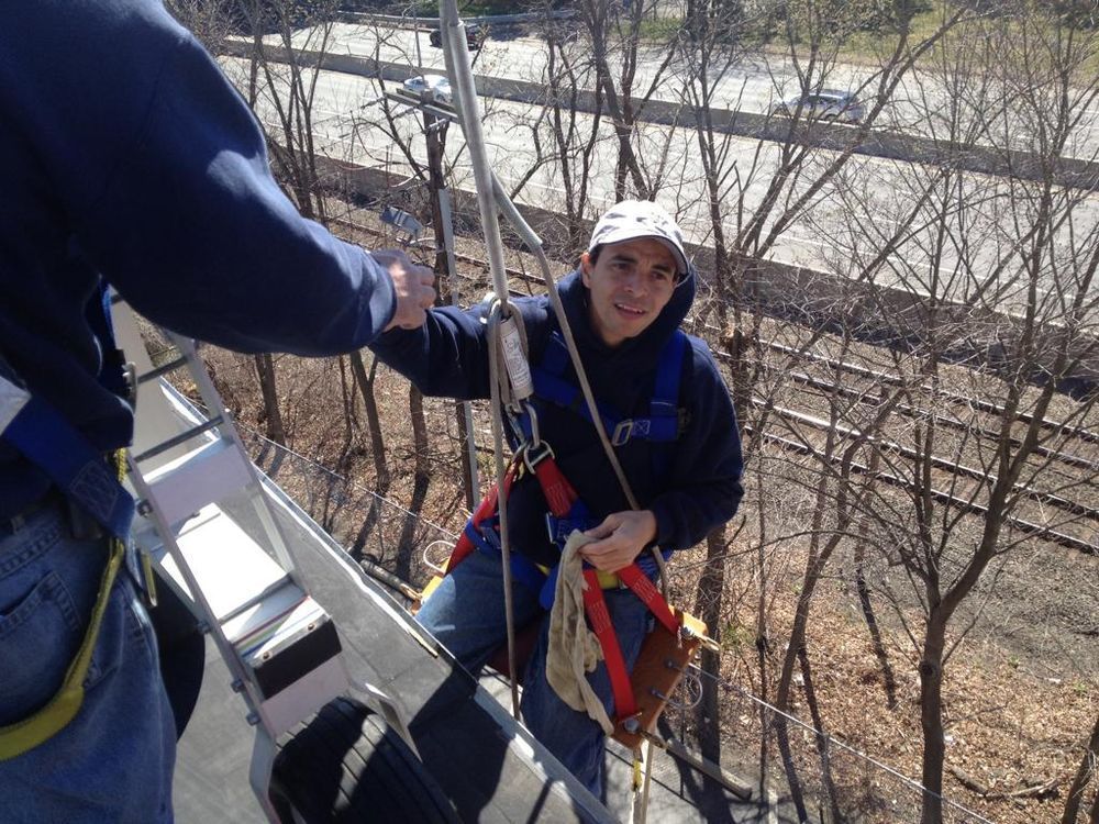 Two climbers on a steep tree lift, one in a helmet and harness reaching up toward another climber.