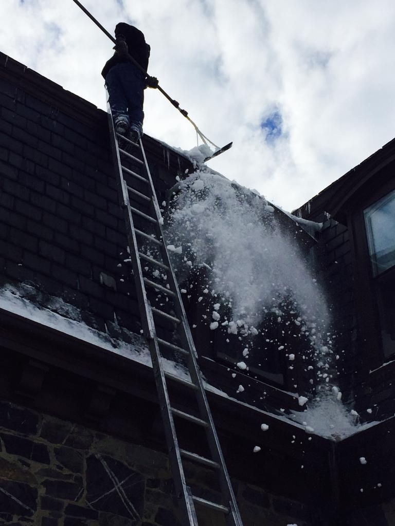 Person on a ladder spraying snow off a rooftop, with snow flying into the air.