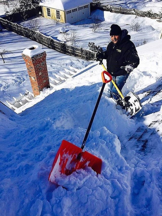 Person shoveling snow from a rooftop with a red snow shovel beside a snow-covered building