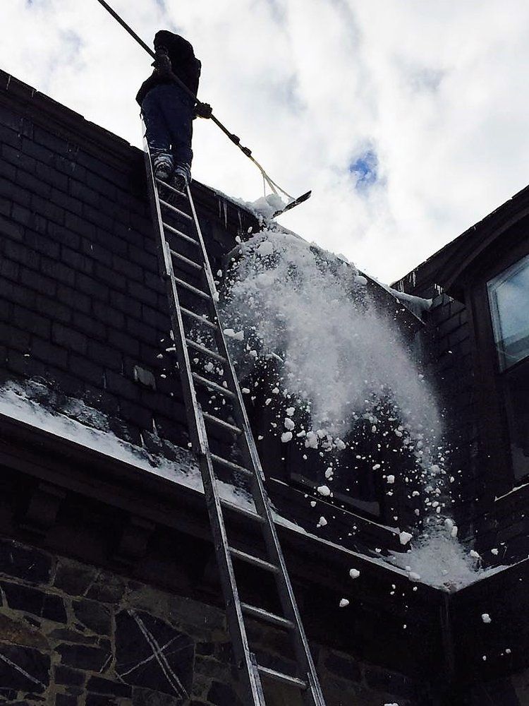 Person on a ladder spraying water from a rooftop onto a smoky, damaged building below