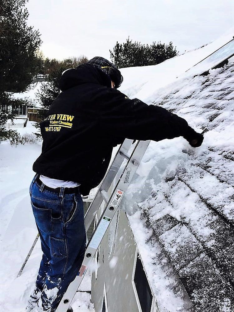 Person in black hoodie clearing snow off a slanted roof with a ladder nearby