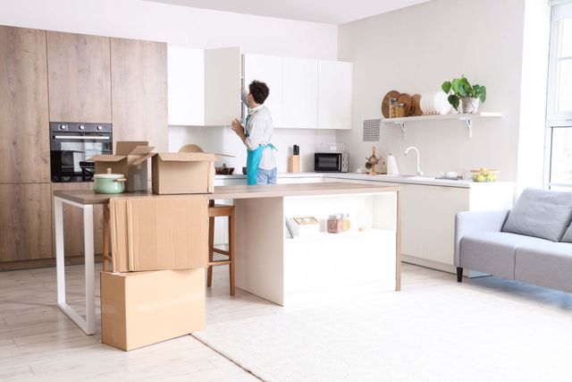 Bright kitchen with person unpacking boxes near a counter and a gray sofa in the background.