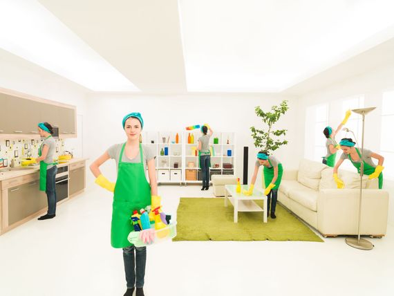 People cleaning a bright living room with brooms, vacuum, and duster, near sofa and rug