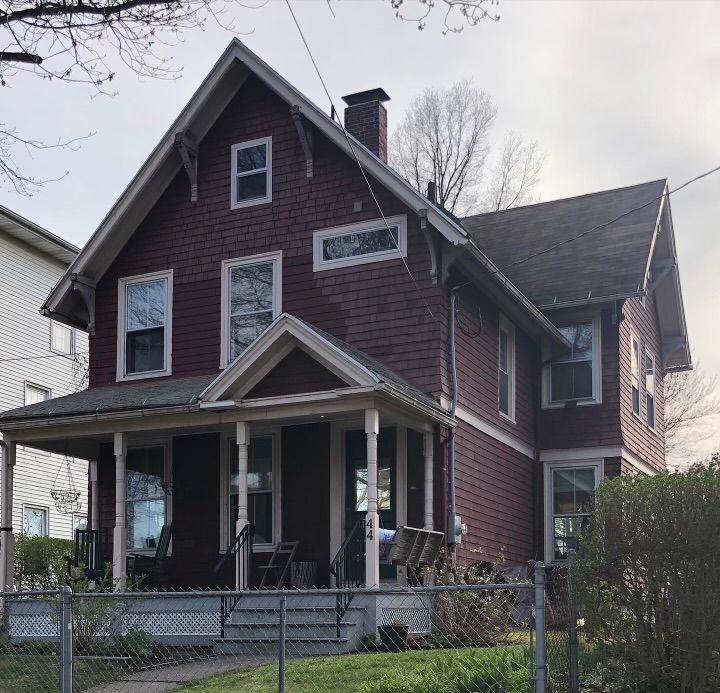 A two-story maroon house with a front porch, visible from behind a chain-link fence on a cloudy day.