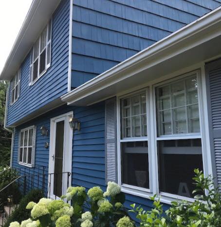 Blue-sided two-story house with white trim, shutters, and windows. White door and hydrangeas in the foreground.
