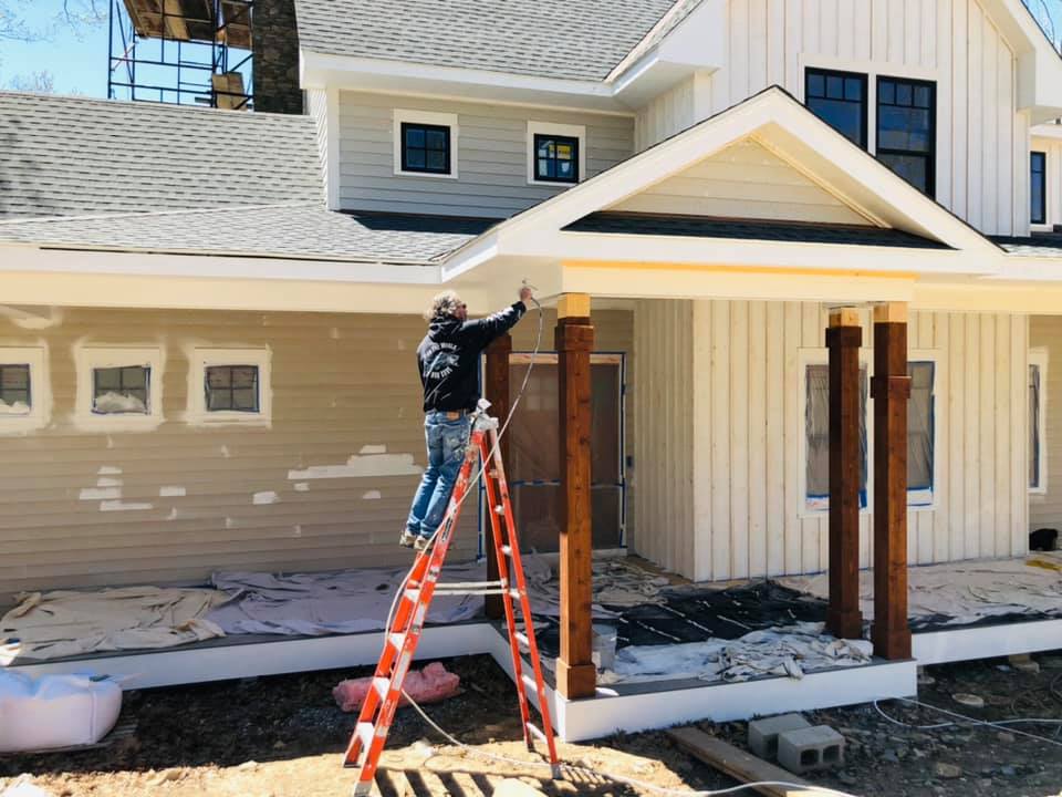Person painting a house exterior on a ladder. The house has white siding, a tan wall, and wooden columns.
