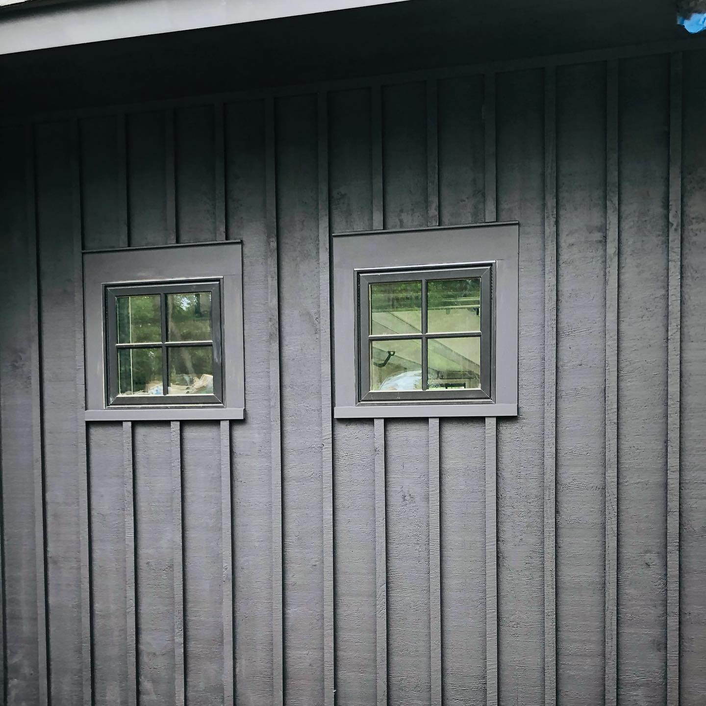 Two square windows with dark frames are set into a vertical, grey-stained wood siding.