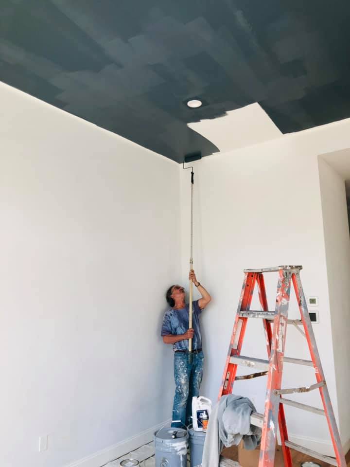 Person painting a dark gray ceiling with a roller, standing on a bucket, with a ladder nearby, in a room with white walls.