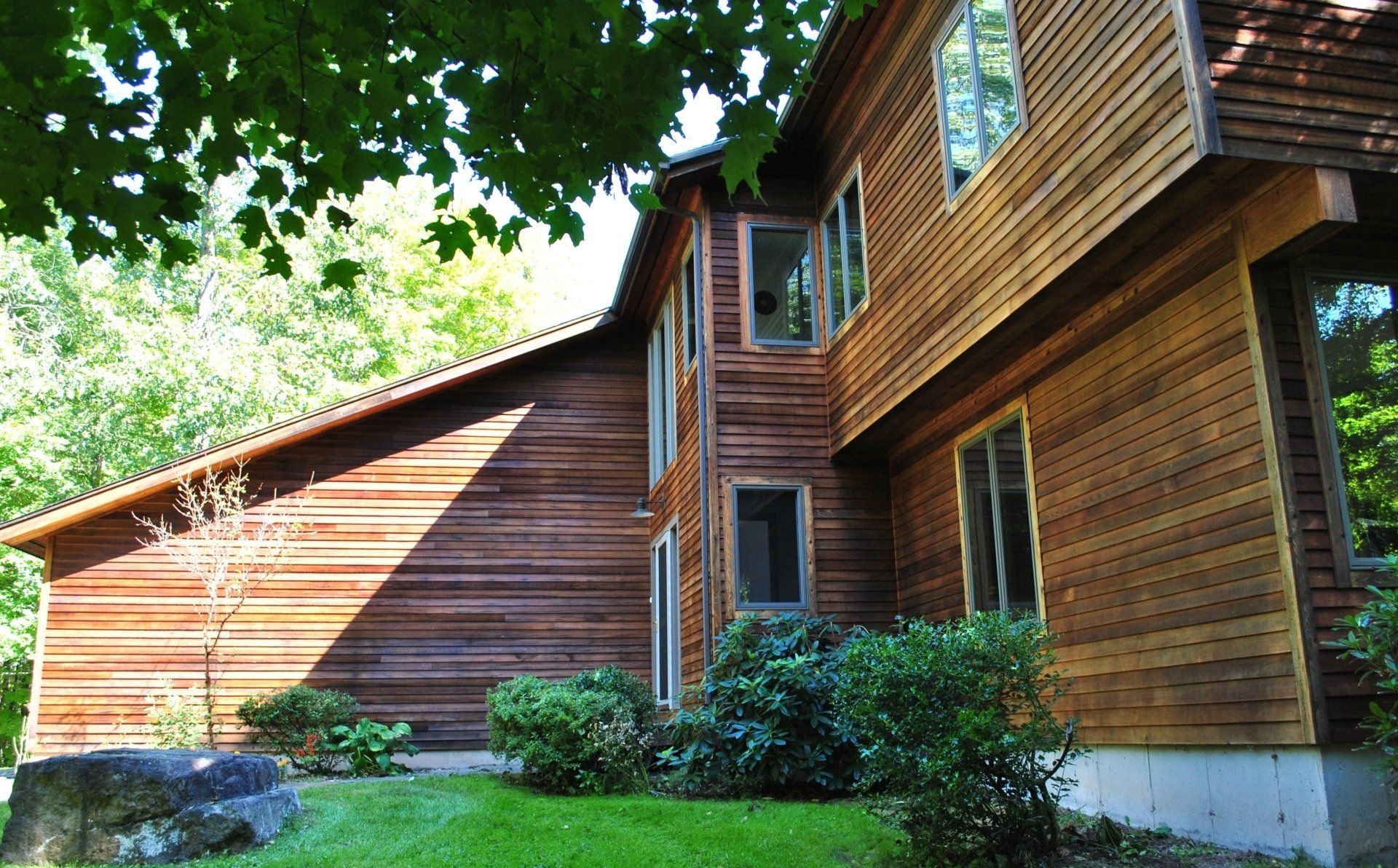 Wooden house with multiple windows and a green lawn, partly shaded by trees.