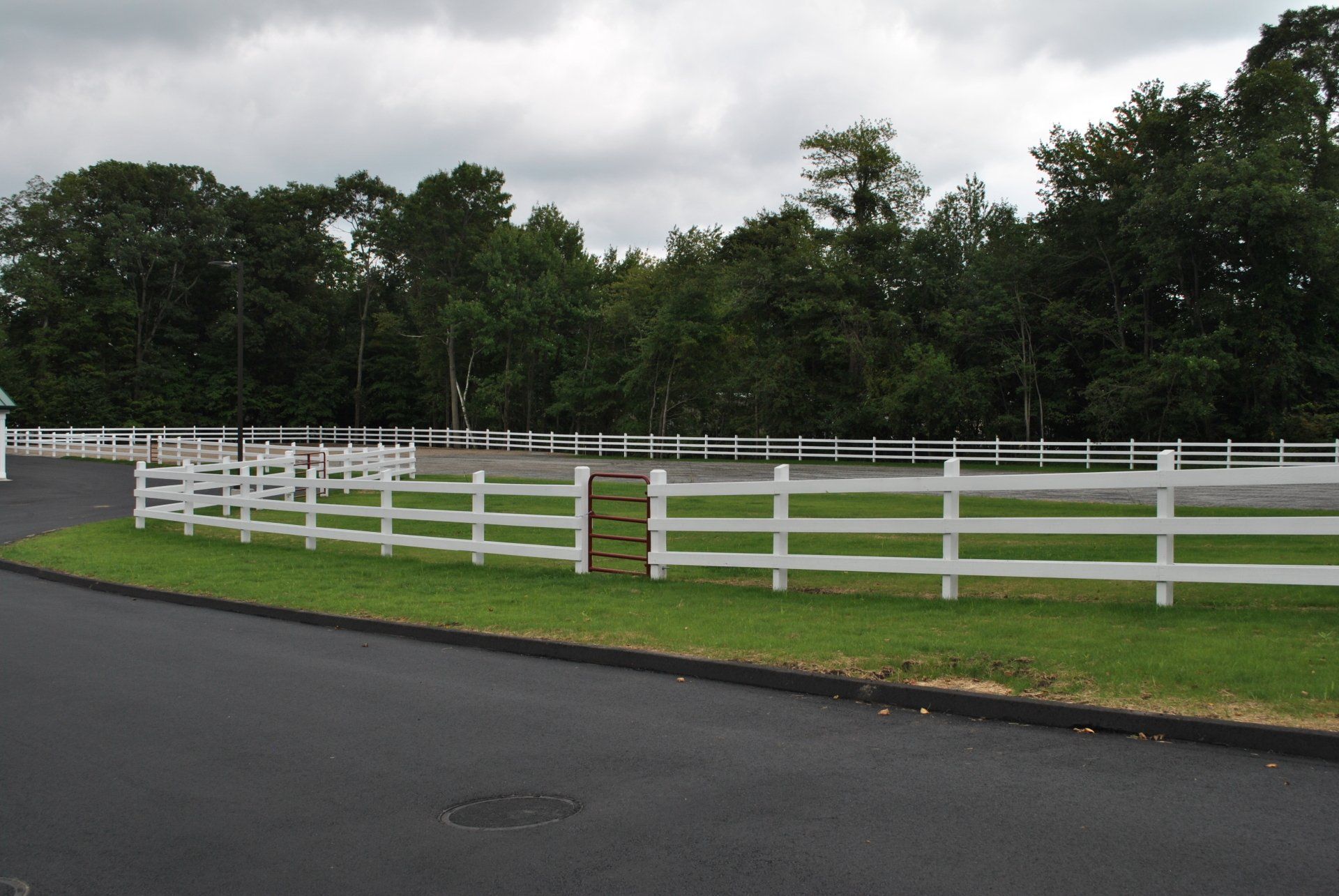 White-fenced horse arena with a paved road in the foreground, lush green grass, and a line of trees under a cloudy sky.