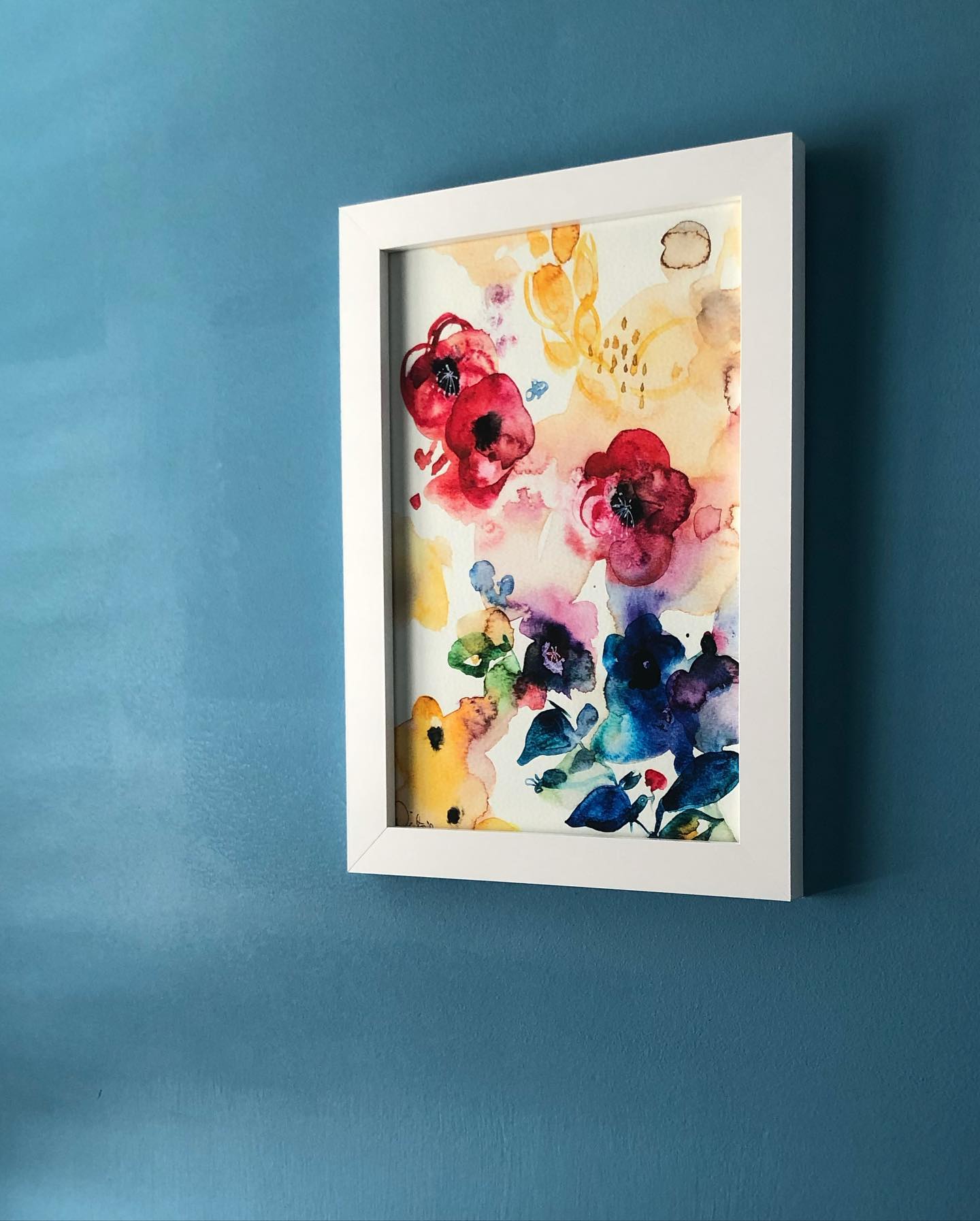 White-framed watercolor floral art against a blue wall. The artwork showcases red and yellow blooms with blue and green accents.
