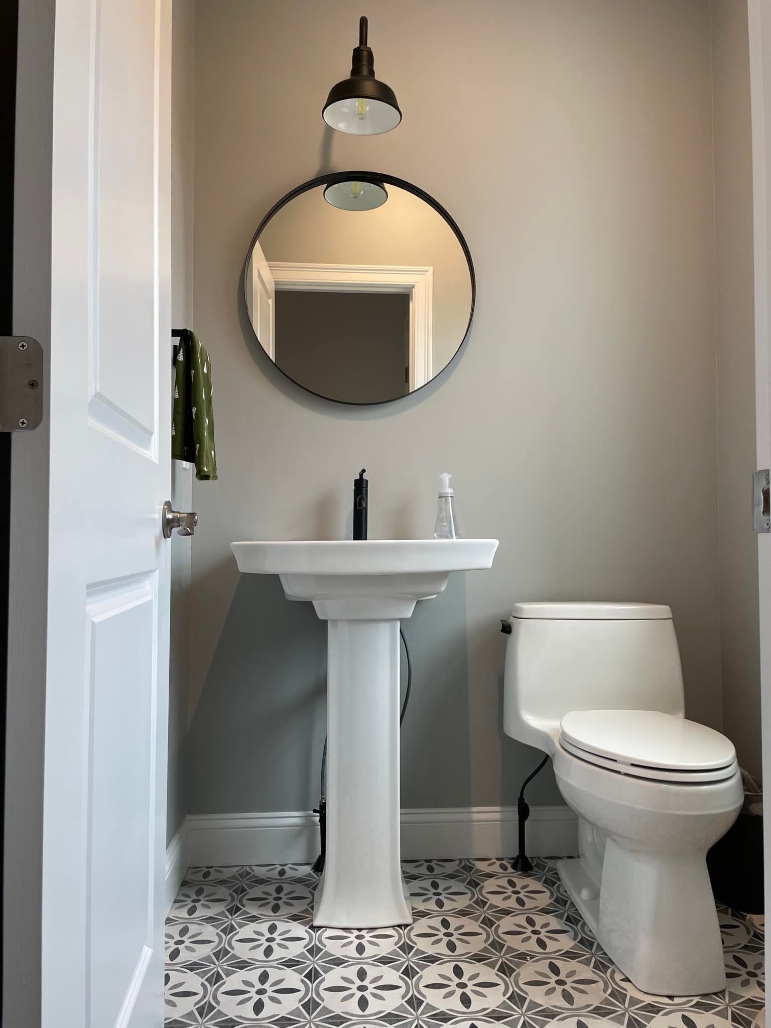 Small powder room with pedestal sink, round mirror, and patterned tile floor. Neutral walls, white trim, and a toilet on the right.