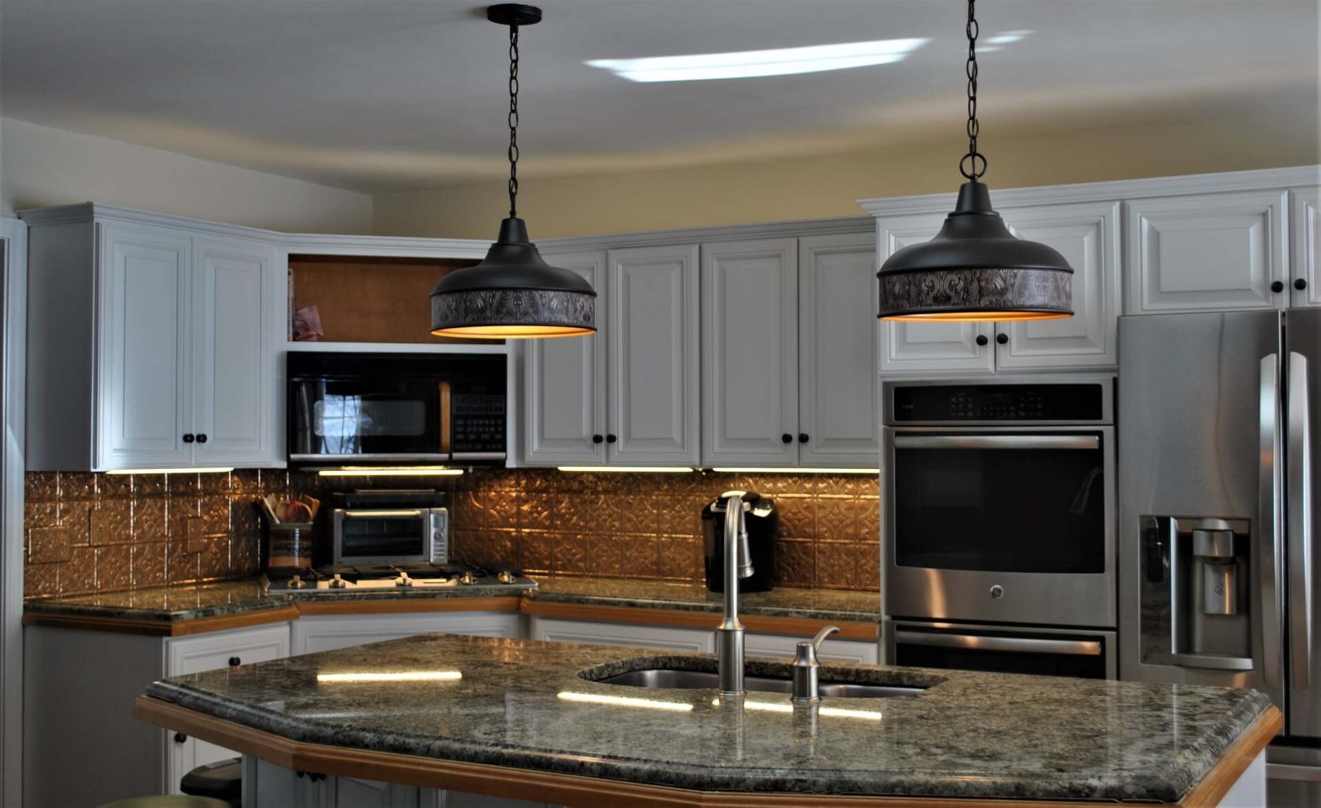 Kitchen with white cabinets, granite countertops, and two pendant lights over an island. Stainless steel appliances and a brown backsplash are visible.