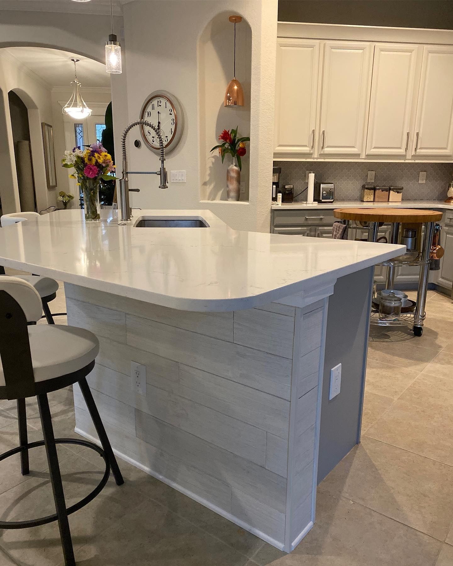 Kitchen island with a light gray tiled finish and white countertops.  White cabinets and stainless steel sink are visible in the background.