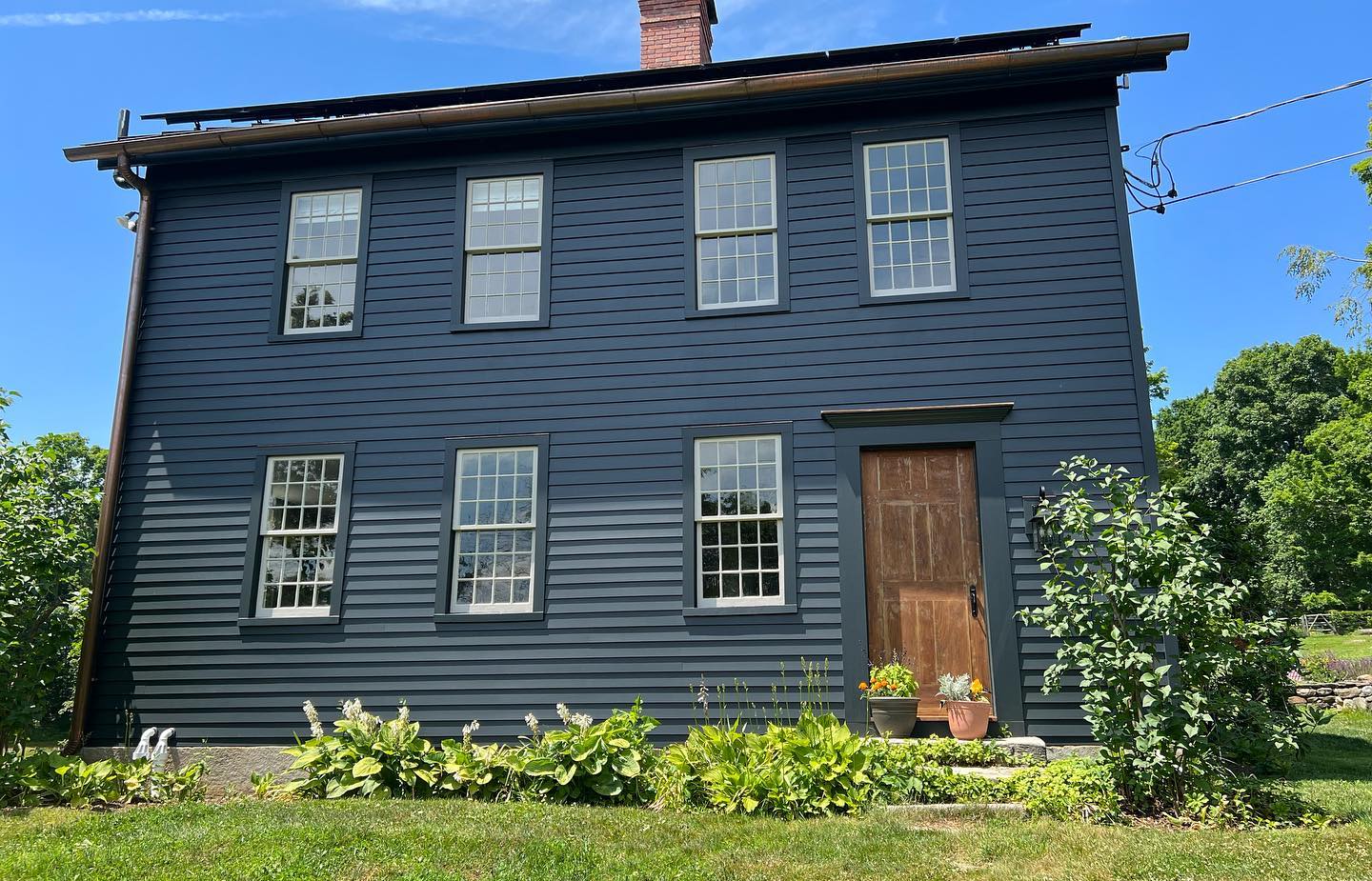 A two-story, dark blue house with six windows and a brown door. The house sits on a grassy lot with some bushes and trees.