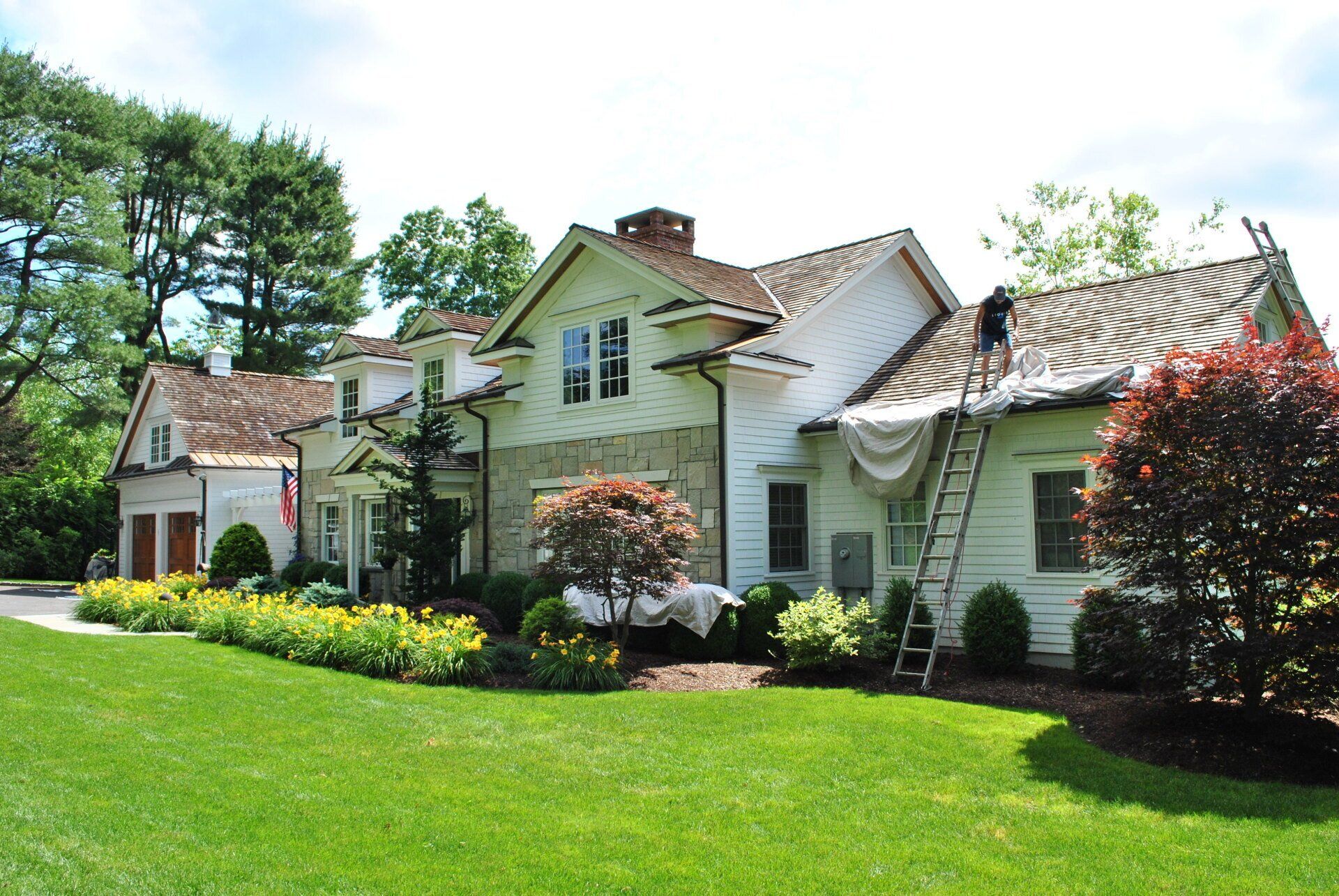 House being renovated; roofers on a ladder, white siding, tan roof, green lawn, and trees in the background.