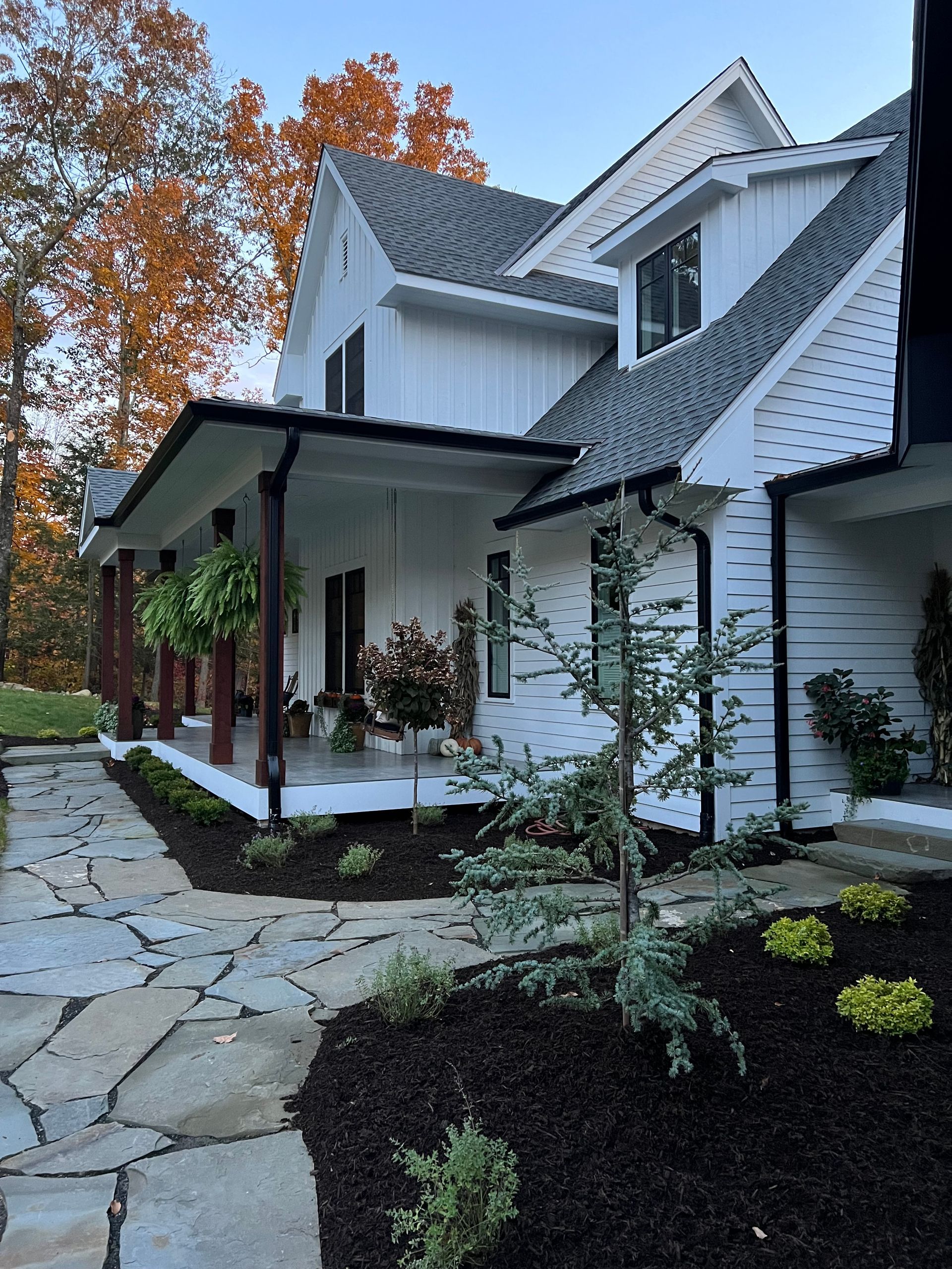 White farmhouse with a wraparound porch, a flagstone path, and a landscaped yard with autumn foliage in the background.
