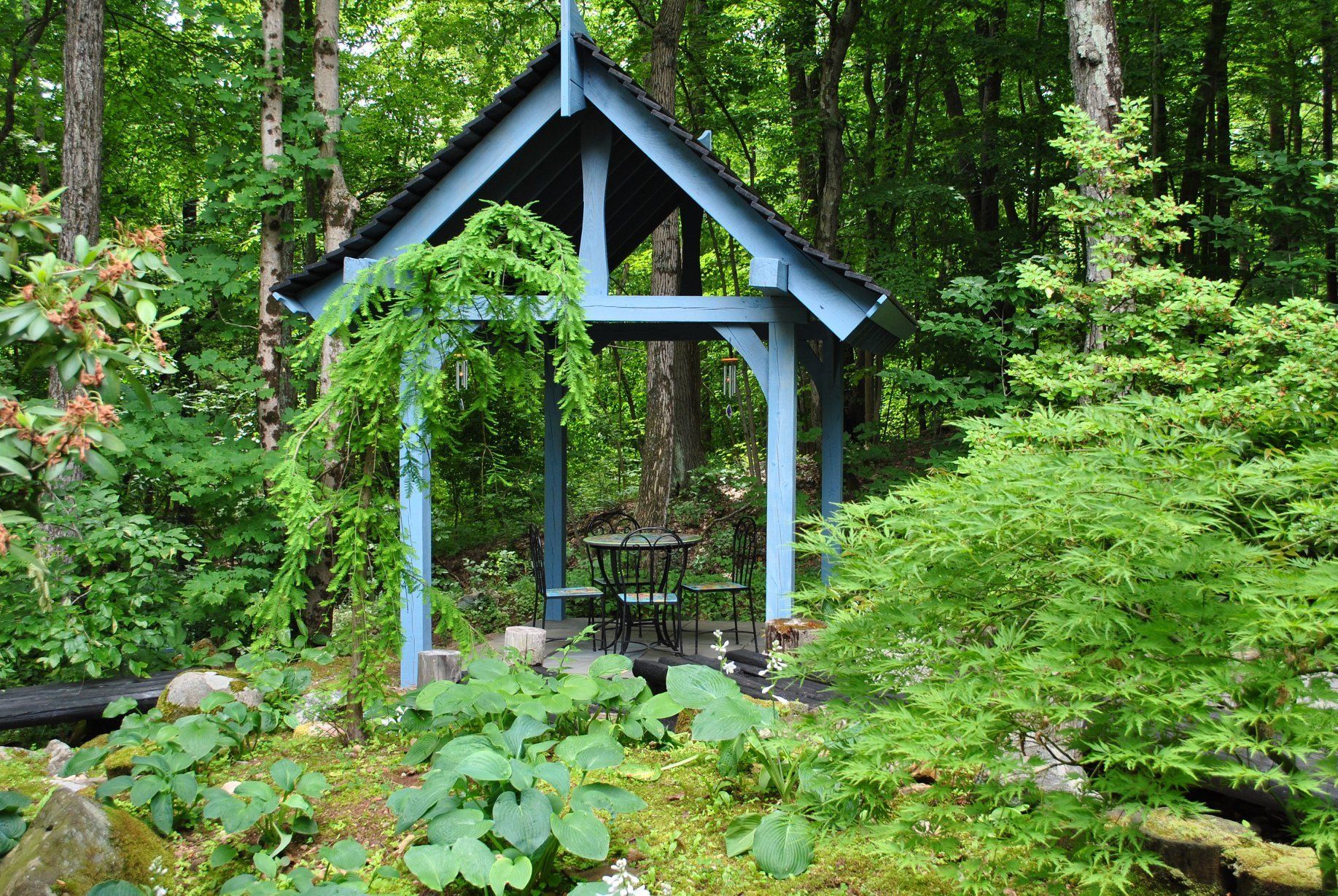 Blue gazebo in a lush green garden, with a table and chairs inside. Vines hang from the gazebo roof.