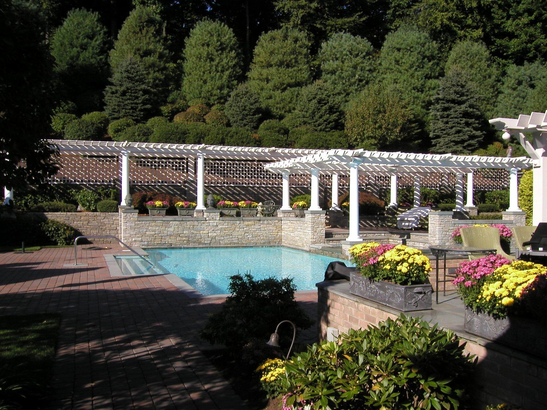 A backyard pool with a white pergola, surrounded by brick and greenery, on a sunny day.