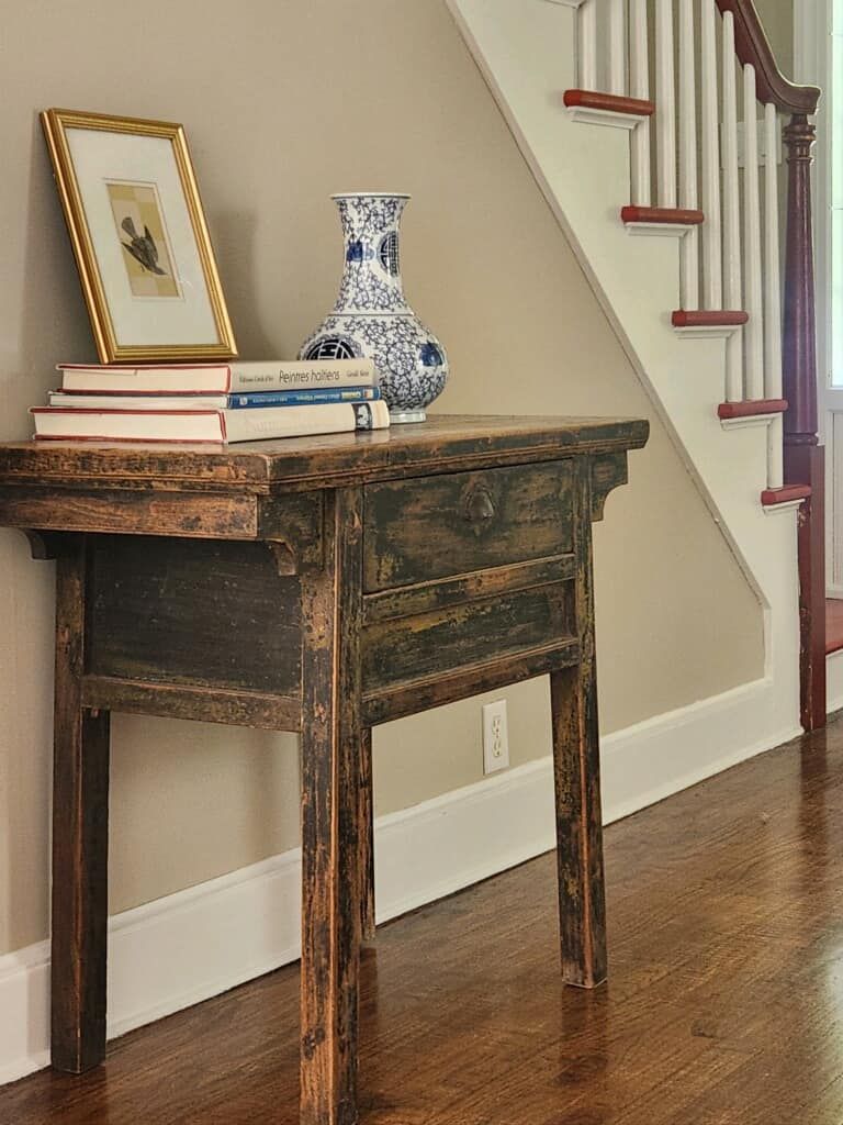 A dark wooden antique table with books, a framed picture, and a blue and white vase in a hallway near stairs.
