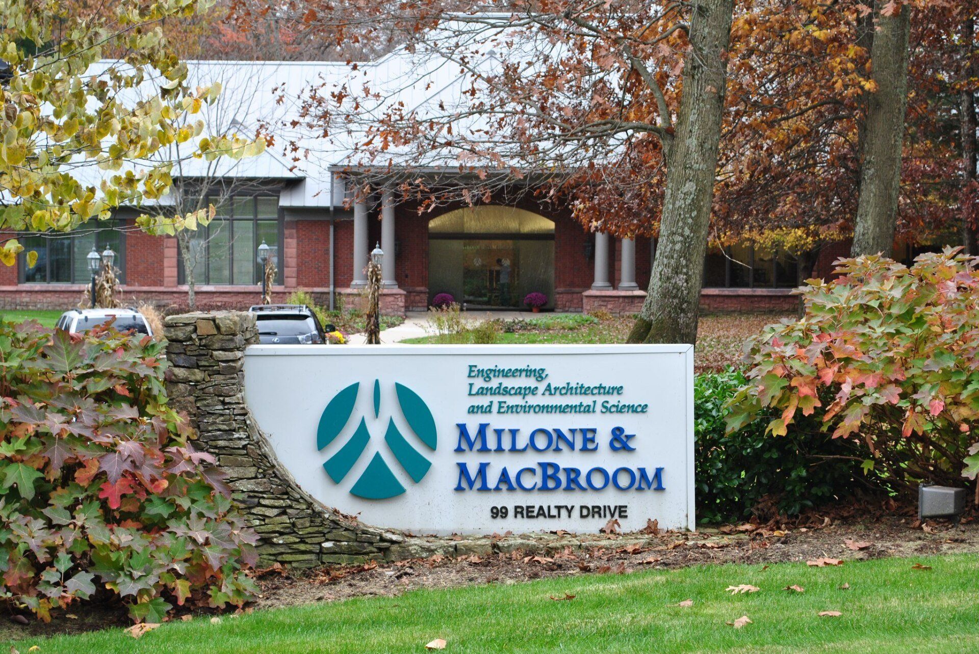 Sign for Milone & MacBroom at 19 Realty Drive. The sign is teal and white, in front of a building with brick and columns.