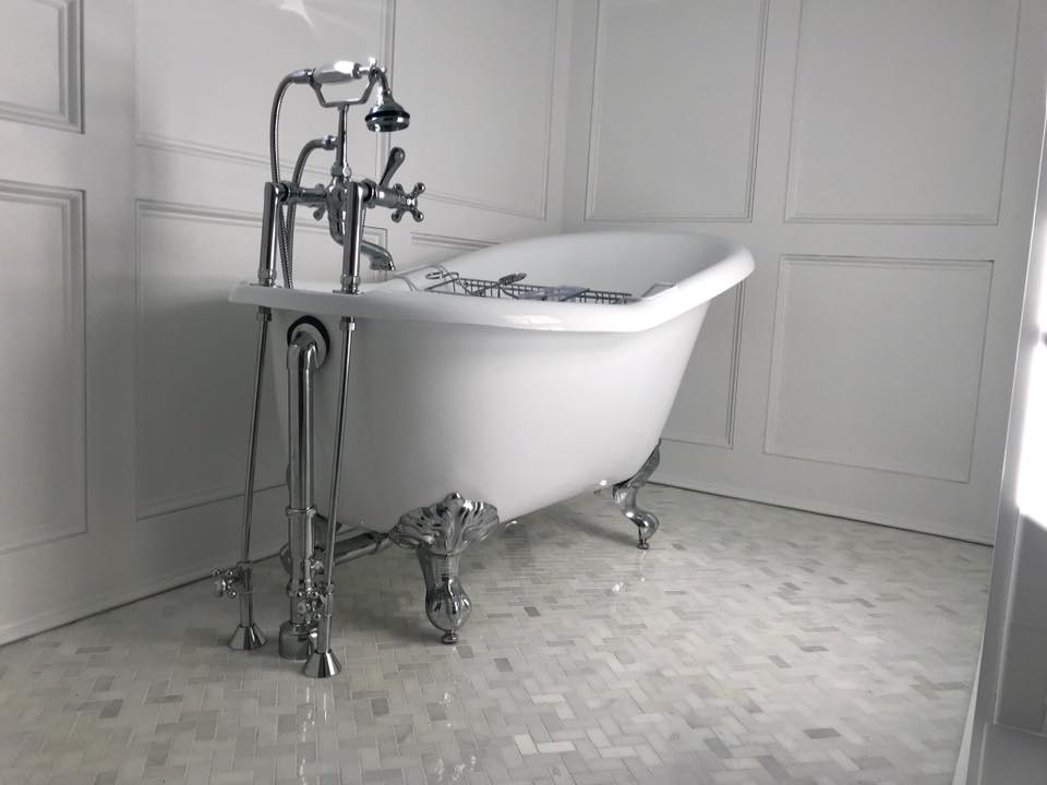 A claw-foot bathtub with ornate chrome fixtures in a white-paneled bathroom with a mosaic tile floor.