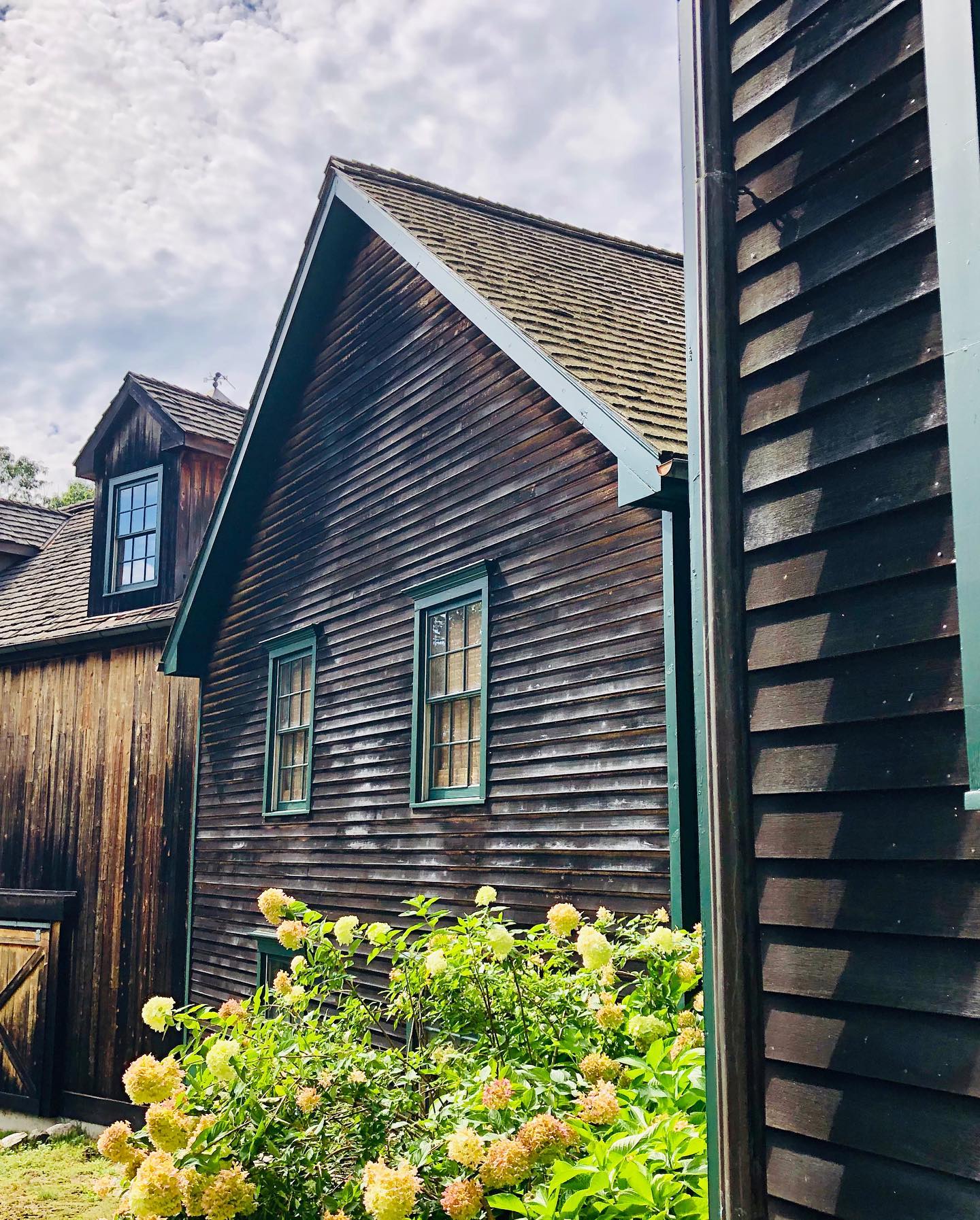 Weathered wooden buildings with green window frames and a dormer window under a cloudy sky. Yellow flowers in the foreground.