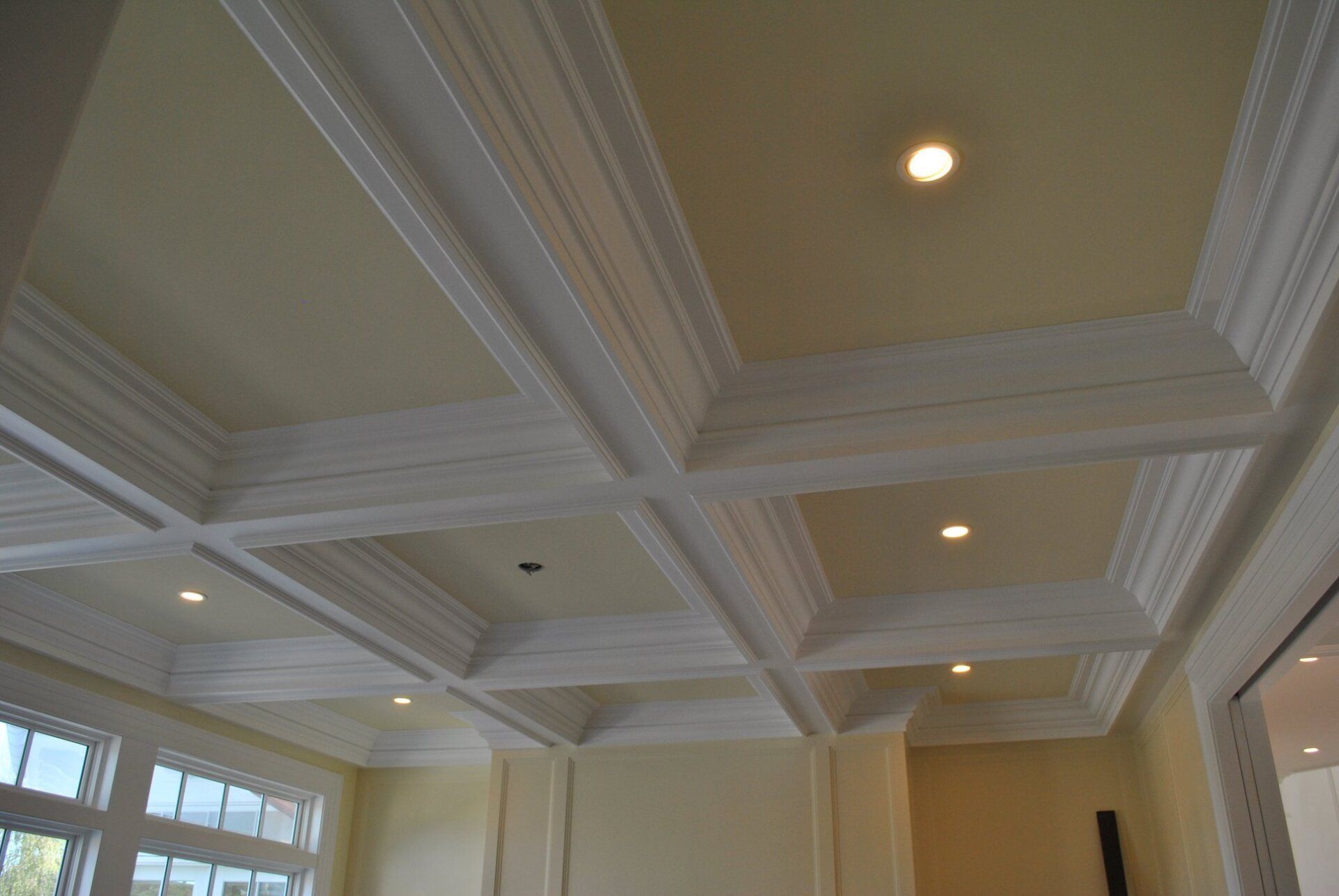 Coffered ceiling with white molding and recessed lights in a room with pale yellow walls.
