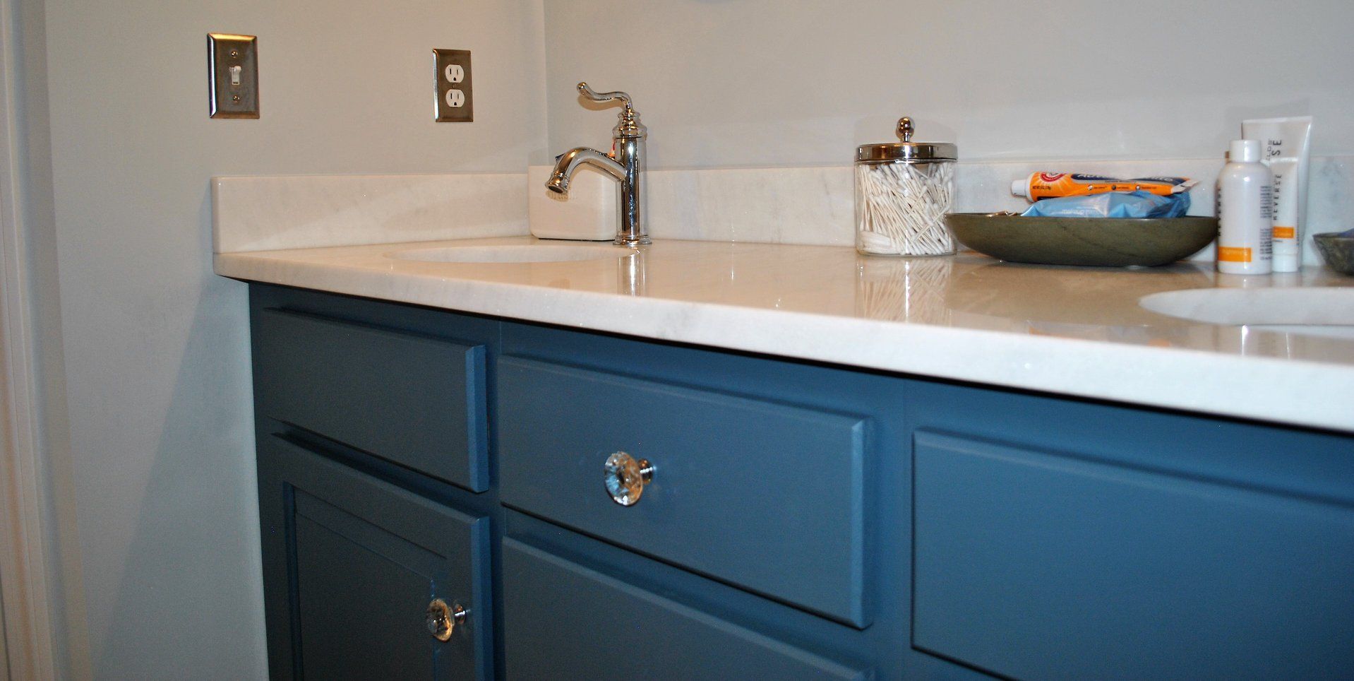 A bathroom vanity with blue cabinets, a white countertop, and chrome fixtures. The counter holds a soap dispenser, bowl, and toiletries.