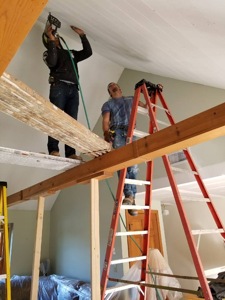 Two men working on a sloped ceiling. One uses a nail gun; the other stands on a ladder, holding tools.