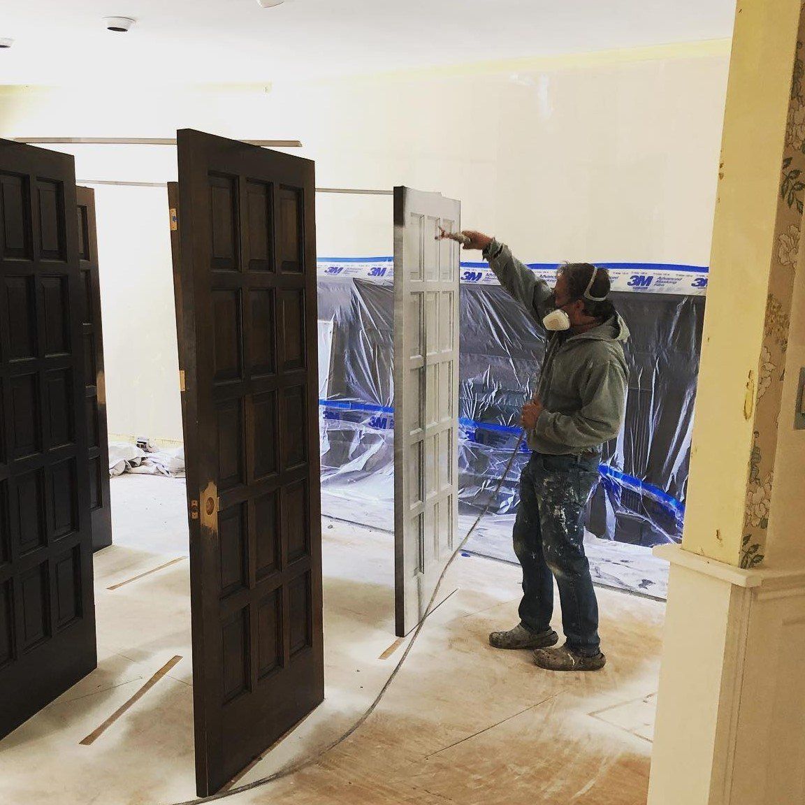 A person sprays paint on a door in an indoor setting. Three other doors stand nearby. The person wears a respirator and work clothes.
