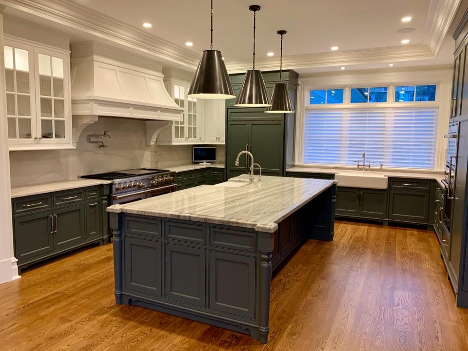 Dark green kitchen with a large island, white countertops, and hanging pendant lights. Wooden floor and a window with white shutters.