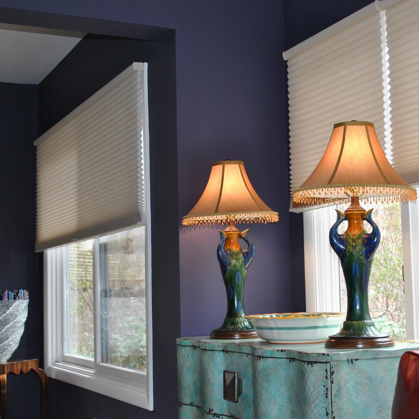 Two lamps with patterned blue bases sit on a teal cabinet against a dark blue wall, near a window with a beige pleated blind.