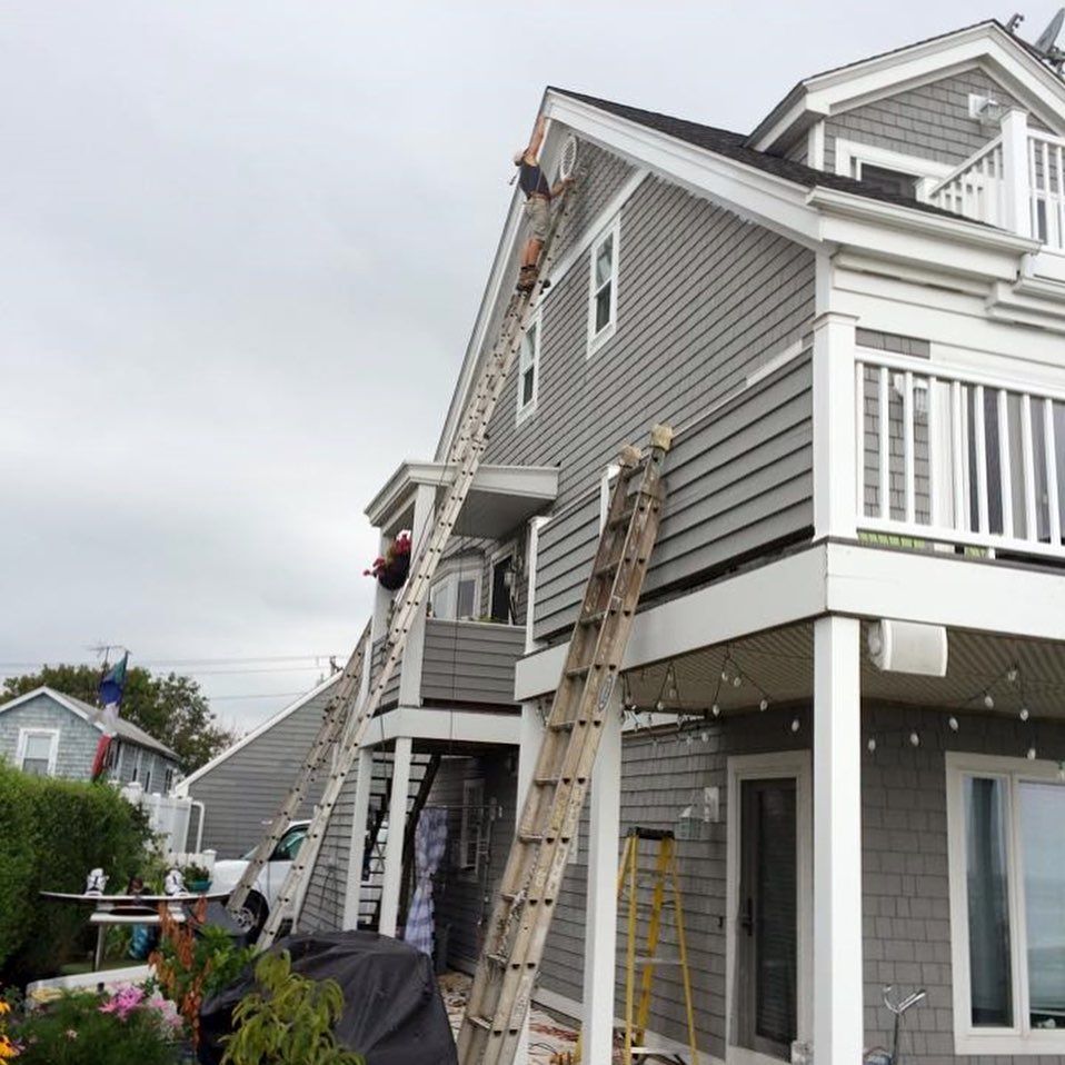 Two workers on tall ladders are repairing the roof of a gray shingled house on a cloudy day.