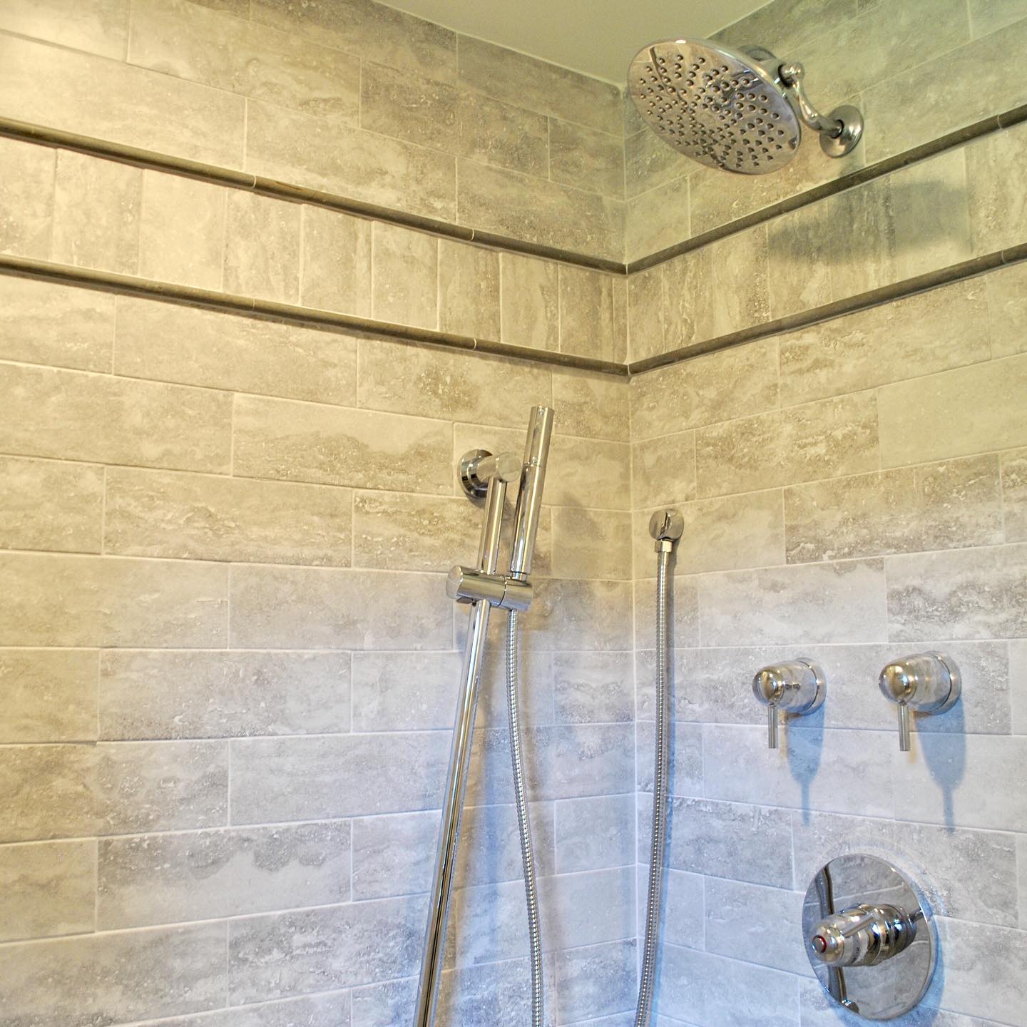 Gray tiled shower with a rain shower head and hand-held shower wand. Chrome fixtures against the neutral walls.