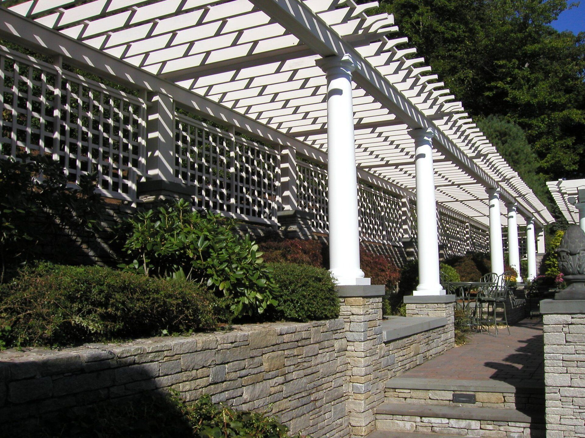 White pergola with lattice and pillars over a stone wall, casting shadows on bushes and stairs.